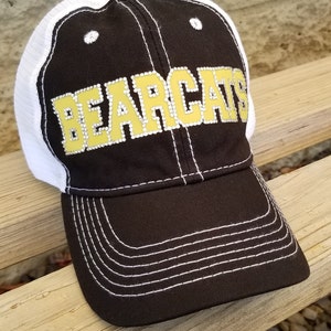 May include: A black baseball cap with a white mesh back and white stitching. The front panel features the word BEARCATS in gold lettering outlined with small clear rhinestones. The cap has a curved brim and a button on the top center.