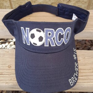 May include: A black visor with the word "NORCO" in silver rhinestones and a white soccer ball with black stitching in the center. The visor has the words "BREWER #15" in silver rhinestones on the side.