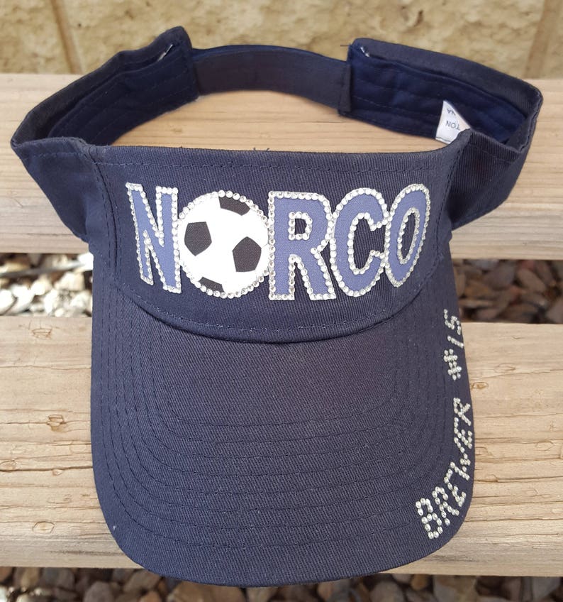 May include: A black visor with the word "NORCO" in silver rhinestones and a white soccer ball with black stitching in the center. The visor has the words "BREWER #15" in silver rhinestones on the side.