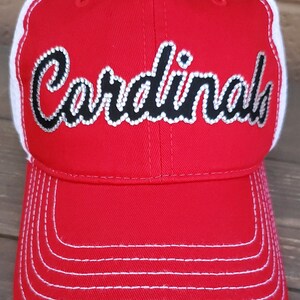 Puede incluir: Gorra de b&eacute;isbol roja y blanca con un logotipo de "Cardinals" en strass negro y blanco.