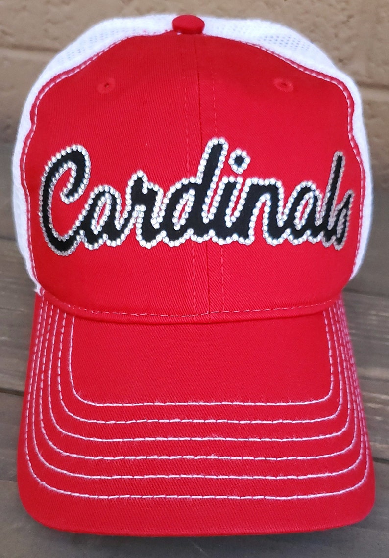 Puede incluir: Gorra de b&eacute;isbol roja y blanca con un logotipo de "Cardinals" en strass negro y blanco.