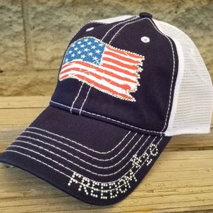 Może przedstawiać: Czapka trucker w kolorze granatowym i białym z nadrukiem zużytej flagi amerykańskiej i napisem "FREEDOM" w cekinach.