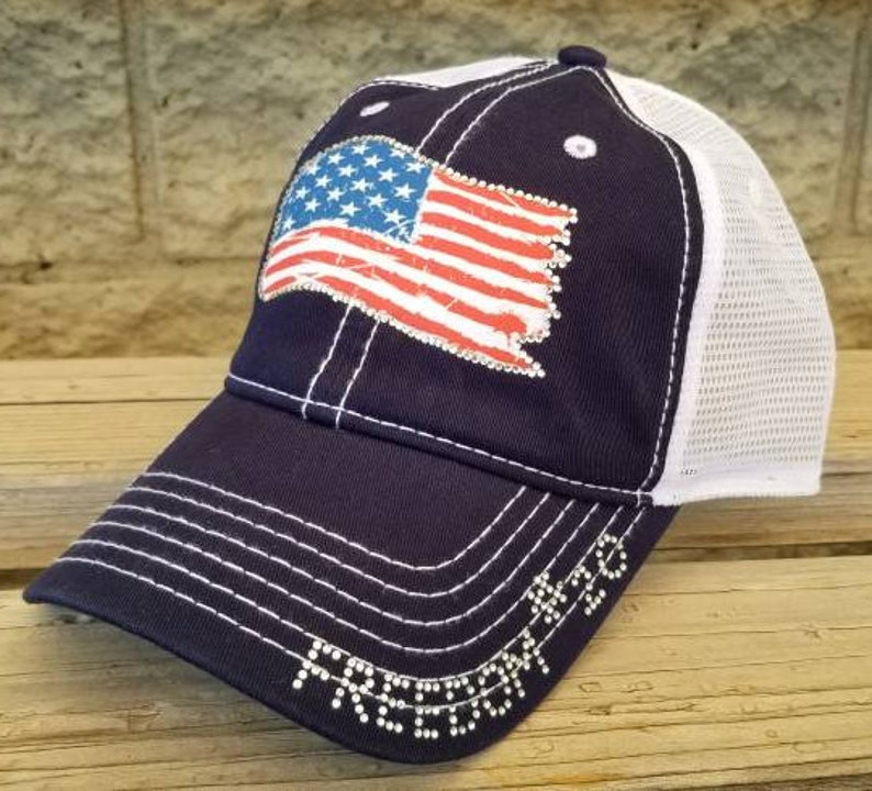 Może przedstawiać: Czapka trucker w kolorze granatowym i białym z nadrukiem zużytej flagi amerykańskiej i napisem "FREEDOM" w cekinach.