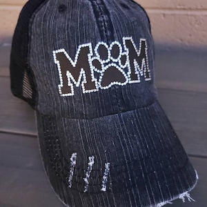May include: Black distressed denim baseball cap with a black and white rhinestone "MOM" design featuring a paw print in the center.