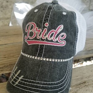 May include: A black and white distressed baseball cap with a white tulle veil. The cap has a pink and white embroidered "Bride" with rhinestones.
