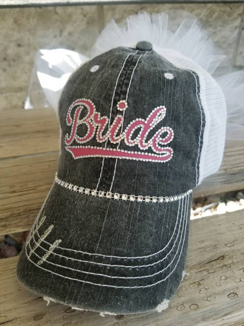May include: A black and white distressed baseball cap with a white tulle veil. The cap has a pink and white embroidered "Bride" with rhinestones.