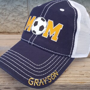 Op de afbeelding: Een donkerblauwe en witte truckercap met een gele en witte voetbal en het woord "MOM" in strass. De pet heeft ook de naam "GRAYSON" in strass op de klep.
