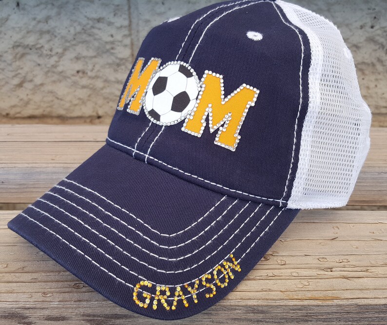 Op de afbeelding: Een donkerblauwe en witte truckercap met een gele en witte voetbal en het woord "MOM" in strass. De pet heeft ook de naam "GRAYSON" in strass op de klep.