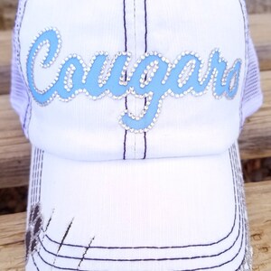 Puede incluir: Gorra de b&eacute;isbol blanca con un dise&ntilde;o "Cougar" azul y blanco con pedrer&iacute;a. La gorra tiene una parte trasera de malla y una visera desgastada.