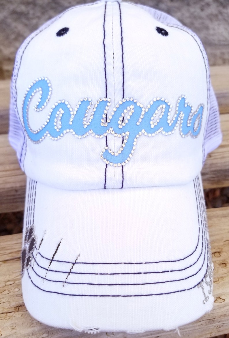 Puede incluir: Gorra de b&eacute;isbol blanca con un dise&ntilde;o "Cougar" azul y blanco con pedrer&iacute;a. La gorra tiene una parte trasera de malla y una visera desgastada.