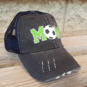 Op de afbeelding: Zwarte en groene truckercap met het woord "MOM" in groene steentjes en een witte voetbal in het midden.