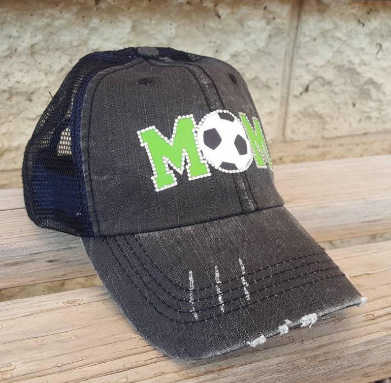 Op de afbeelding: Zwarte en groene truckercap met het woord "MOM" in groene steentjes en een witte voetbal in het midden.