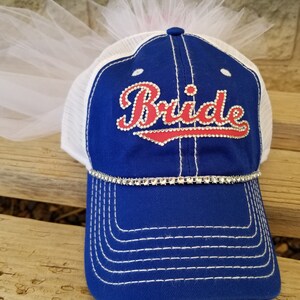 May include: A blue and white baseball cap with a white tulle veil attached to the top. The cap has a red and white rhinestone design that says "Bride".