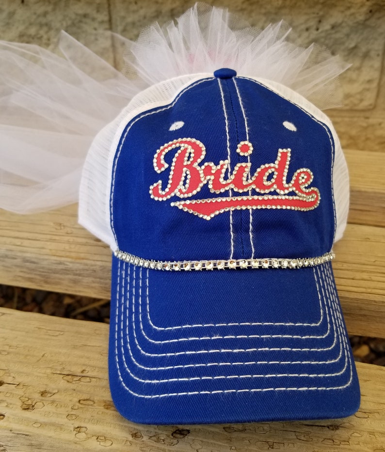 May include: A blue and white baseball cap with a white tulle veil attached to the top. The cap has a red and white rhinestone design that says "Bride".