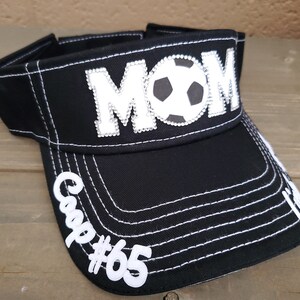 May include: Black visor with white stitching and a white "MOM" with rhinestones and a soccer ball in the center. The visor has a white "Cap #65" on the side.