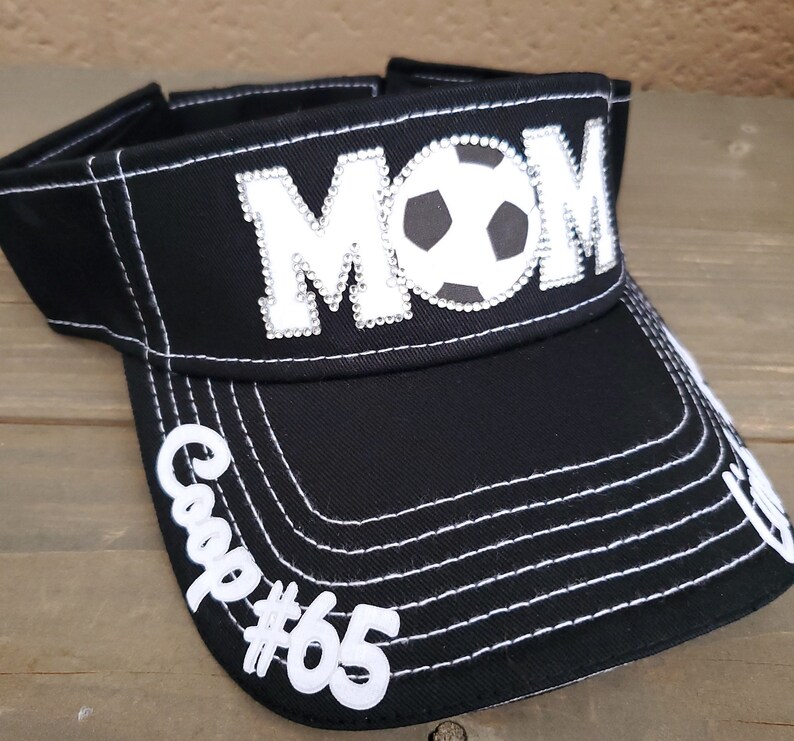 May include: Black visor with white stitching and a white "MOM" with rhinestones and a soccer ball in the center. The visor has a white "Cap #65" on the side.