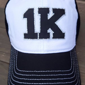 May include: A white and black baseball cap with a black "1K" design on the front. The "1K" is covered in rhinestones. The cap has a black mesh back and a black brim with white stitching.