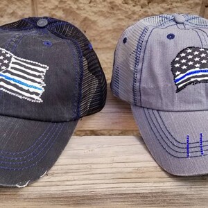 May include: Two distressed baseball caps with a thin blue line American flag design. The caps are black and grey with black mesh back panels.
