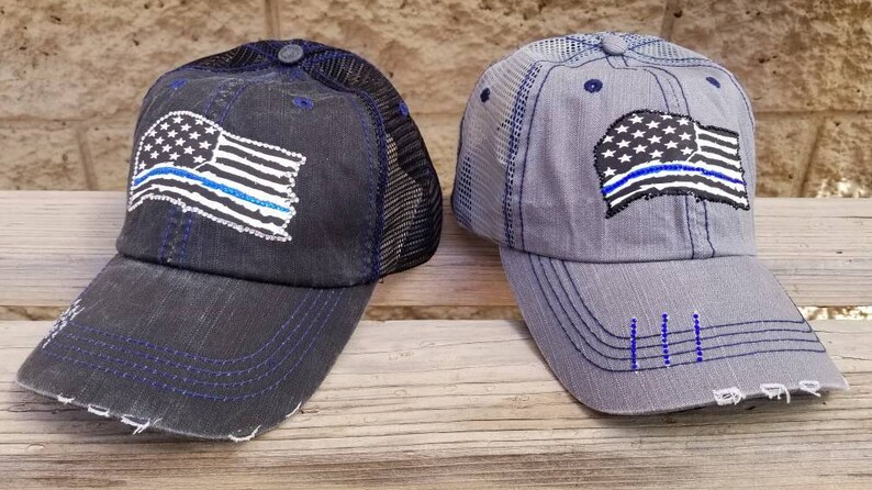 May include: Two distressed baseball caps with a thin blue line American flag design. The caps are black and grey with black mesh back panels.