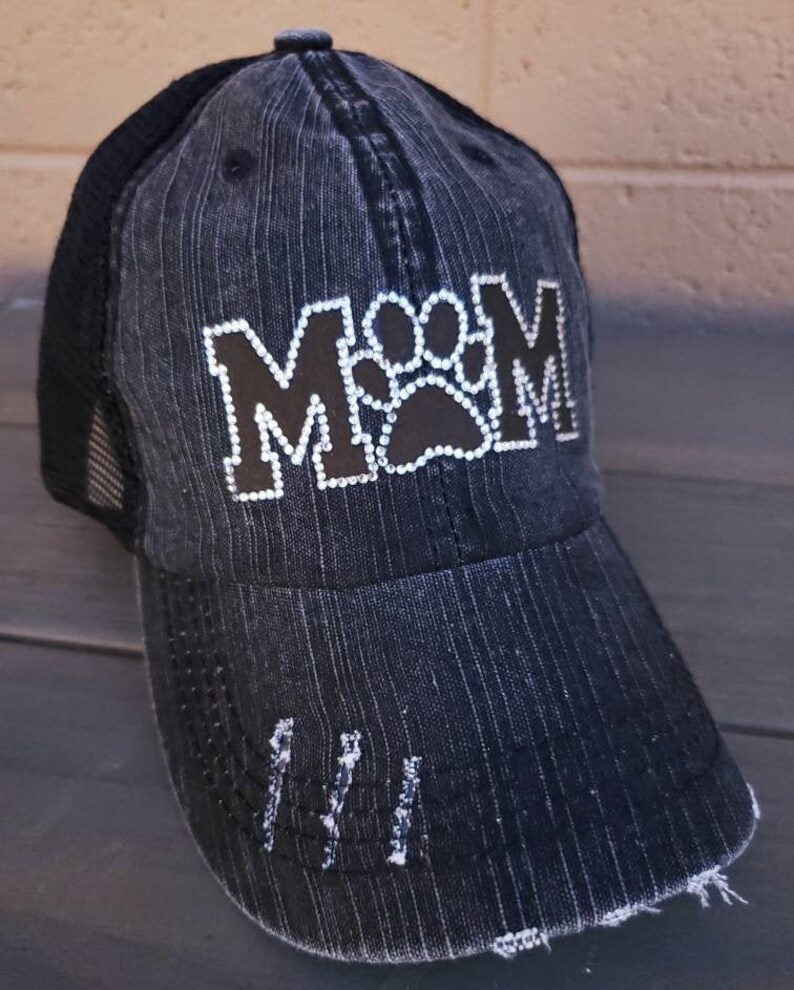 May include: Black and grey distressed baseball cap with a black paw print and the word "MOM" in rhinestones.