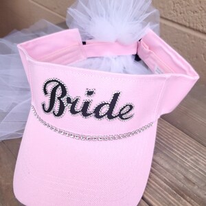 K&ouml;nnte beinhalten: Rosa Visier mit einem wei&szlig;en T&uuml;llschleier und dem Wort "Bride" in Schwarz mit Strasssteinen.