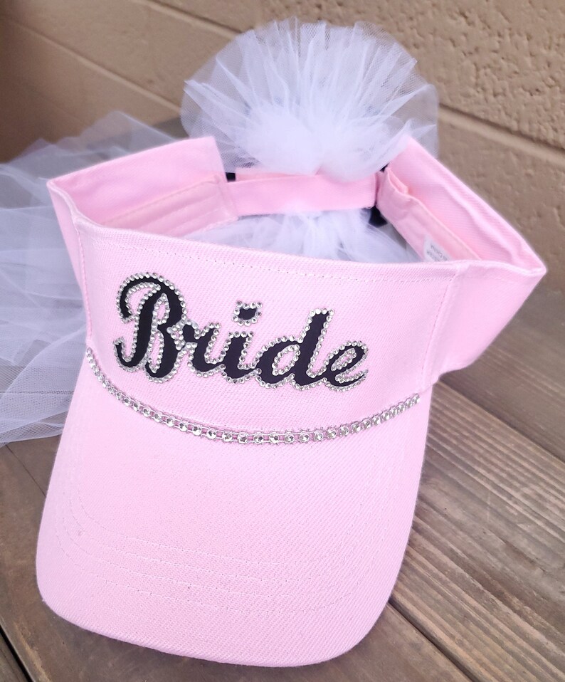 K&ouml;nnte beinhalten: Rosa Visier mit einem wei&szlig;en T&uuml;llschleier und dem Wort "Bride" in Schwarz mit Strasssteinen.