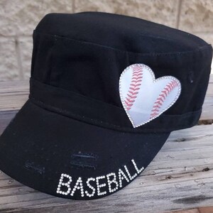 Pode incluir: Bon&eacute; de beisebol preto em estilo militar com um design de beisebol em forma de cora&ccedil;&atilde;o. O cora&ccedil;&atilde;o de beisebol &eacute; branco com costuras vermelhas e uma borda de strass. A palavra "BASEBALL" est&aacute; escrita com strass na aba.