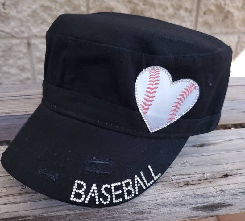 Pode incluir: Bon&eacute; de beisebol preto em estilo militar com um design de beisebol em forma de cora&ccedil;&atilde;o. O cora&ccedil;&atilde;o de beisebol &eacute; branco com costuras vermelhas e uma borda de strass. A palavra "BASEBALL" est&aacute; escrita com strass na aba.