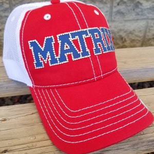 May include: A red baseball cap with a white mesh back and white stitching. The front features the word MATRIX in blue block letters outlined with small clear rhinestones. The cap has a red button on top and white eyelets on the red front panels.
