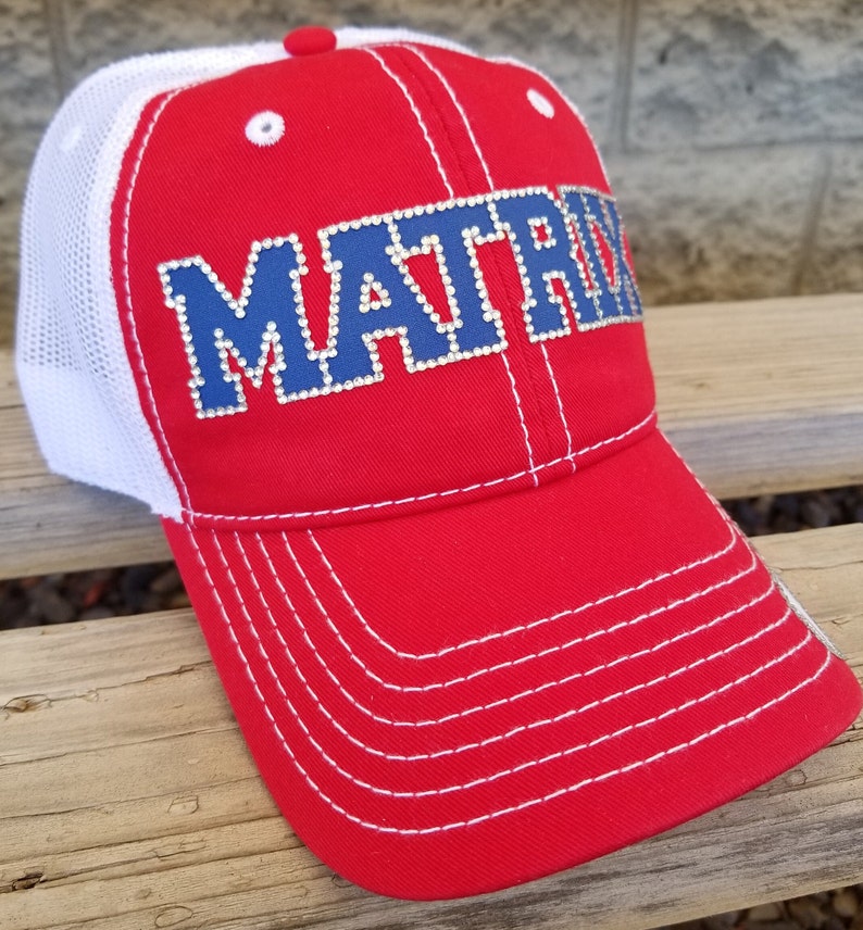 May include: A red baseball cap with a white mesh back and white stitching. The front features the word MATRIX in blue block letters outlined with small clear rhinestones. The cap has a red button on top and white eyelets on the red front panels.