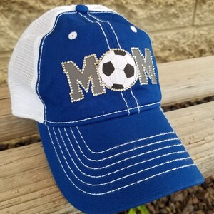 Op de afbeelding: Blauw-witte baseballpet met een mesh achterkant. De voorkant van de pet heeft een grijze en witte voetbal met het woord "MOM" in strass.