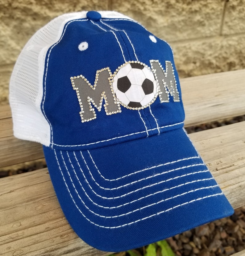 Op de afbeelding: Blauw-witte baseballpet met een mesh achterkant. De voorkant van de pet heeft een grijze en witte voetbal met het woord "MOM" in strass.