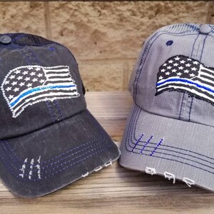 May include: Two black and grey trucker hats with a distressed look. Both hats feature a white American flag with a blue line across it. The blue line represents support for law enforcement.