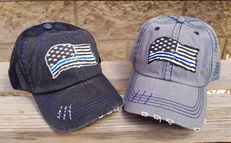 May include: Two black and grey trucker hats with a distressed look. Both hats feature a white American flag with a blue line across it. The blue line represents support for law enforcement.