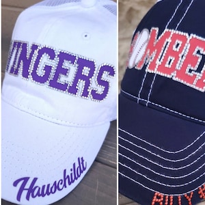 May include: Two baseball caps with rhinestones. The white cap has purple rhinestones spelling "RANGERS" and "Hauschildt". The navy blue cap has red rhinestones spelling "BOMBERS" and "BILLY #".