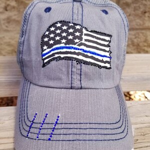 May include: A grey and blue baseball cap with a distressed look. The cap features a black and white American flag with a blue line across it. The flag is embellished with rhinestones.
