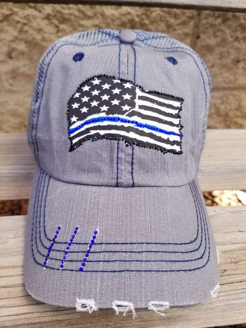 May include: A grey and blue baseball cap with a distressed look. The cap features a black and white American flag with a blue line across it. The flag is embellished with rhinestones.