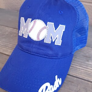 May include: A royal blue baseball cap with a white baseball and the word "MOM" in rhinestones. The word "Babe" is written in white on the brim of the cap.