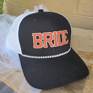 May include: A black and white trucker hat featuring the word BRIDE in orange letters with rhinestone borders. A single row of rhinestones accents the base of the front panel, and a white tulle veil is attached to the back of the hat.