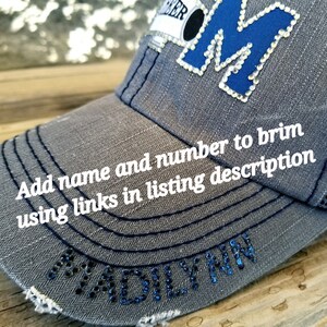 Op de afbeelding: Grijze baseballpet met een blauwe en witte letter M met strasssteentjes aan de voorkant. De rand van de pet heeft een zwarte bies en de woorden "Voeg naam en nummer toe aan de rand met behulp van links in de productbeschrijving" staan op de pet.