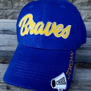 May include: Blue baseball cap with the word "Braves" in yellow with rhinestones. The cap has a white patch with a megaphone and the word "Cheer" in rhinestones. The side of the cap has the word "Jordan" in rhinestones.