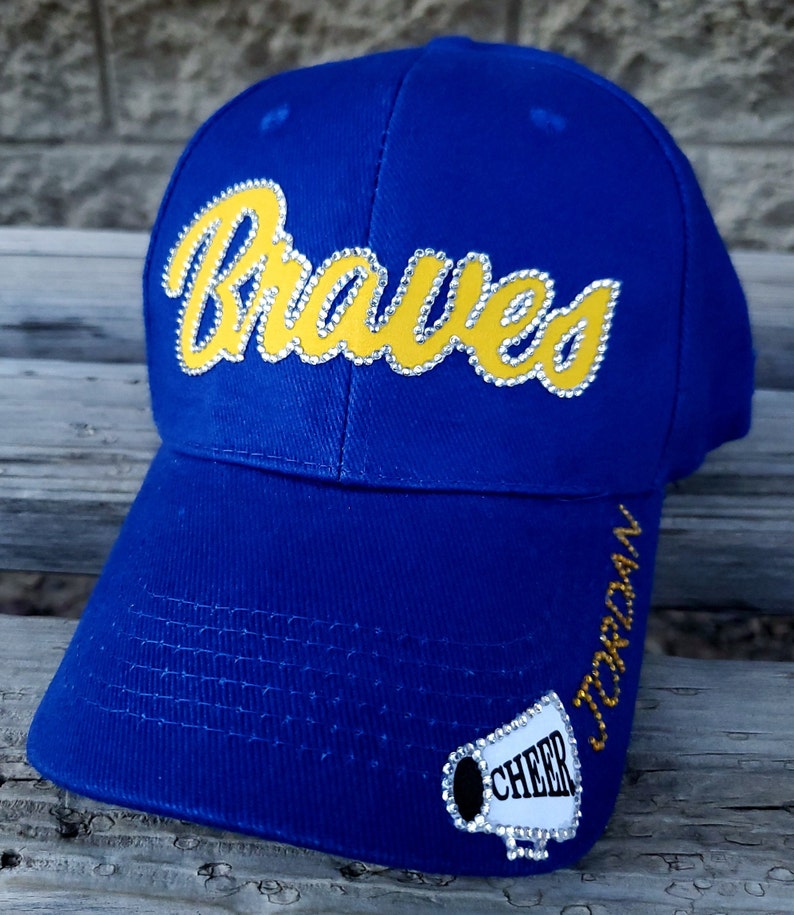 May include: Blue baseball cap with the word "Braves" in yellow with rhinestones. The cap has a white patch with a megaphone and the word "Cheer" in rhinestones. The side of the cap has the word "Jordan" in rhinestones.