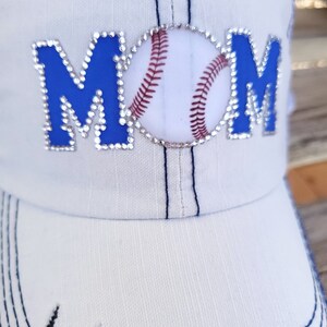 May include: White baseball cap with blue and rhinestone lettering that says "MOM" with a baseball graphic in the center.