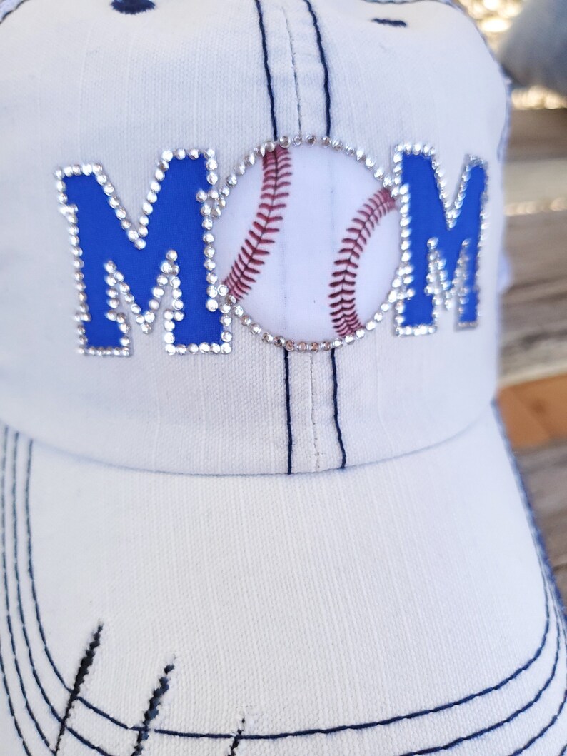 May include: White baseball cap with blue and rhinestone lettering that says "MOM" with a baseball graphic in the center.