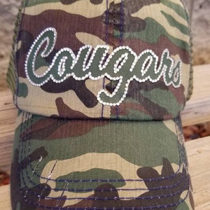 May include: Camouflage trucker hat with a green and white "Cougars" design with rhinestones. The hat has a mesh back and a distressed brim.