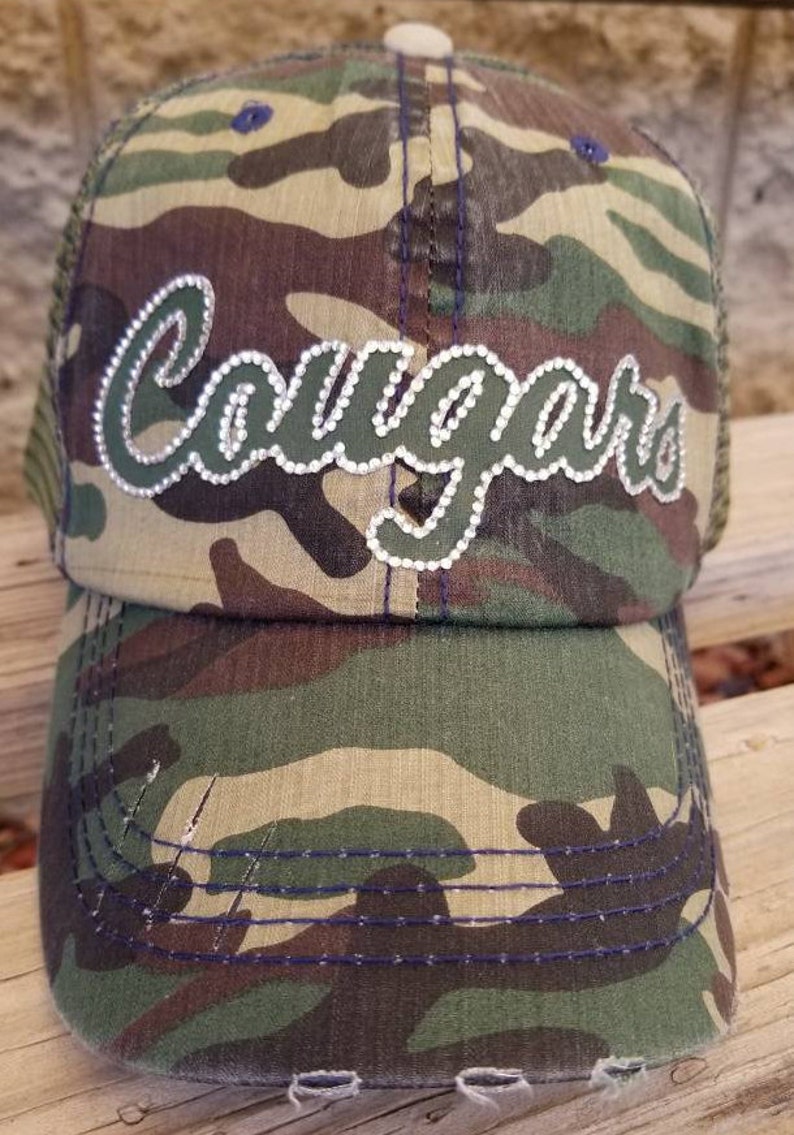 May include: Camouflage trucker hat with a green and white "Cougars" design with rhinestones. The hat has a mesh back and a distressed brim.