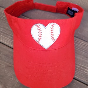 May include: A bright red visor featuring a central heart-shaped patch designed to look like a white baseball with red stitching. The heart patch is outlined with small, clear, reflective rhinestones. The visor has an adjustable strap at the back.