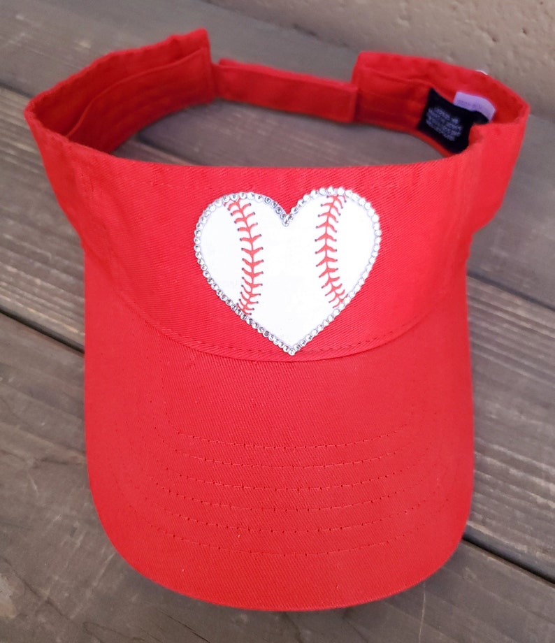 May include: A bright red visor featuring a central heart-shaped patch designed to look like a white baseball with red stitching. The heart patch is outlined with small, clear, reflective rhinestones. The visor has an adjustable strap at the back.