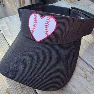 May include: A black visor featuring a white heart-shaped patch on the front panel. The heart patch displays a red baseball stitch pattern across its center. The visor has a structured brim and an adjustable strap closure at the back for a secure fit.