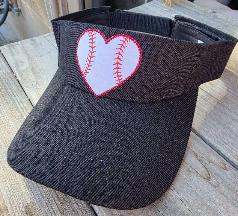 May include: A black visor featuring a white heart-shaped patch on the front panel. The heart patch displays a red baseball stitch pattern across its center. The visor has a structured brim and an adjustable strap closure at the back for a secure fit.